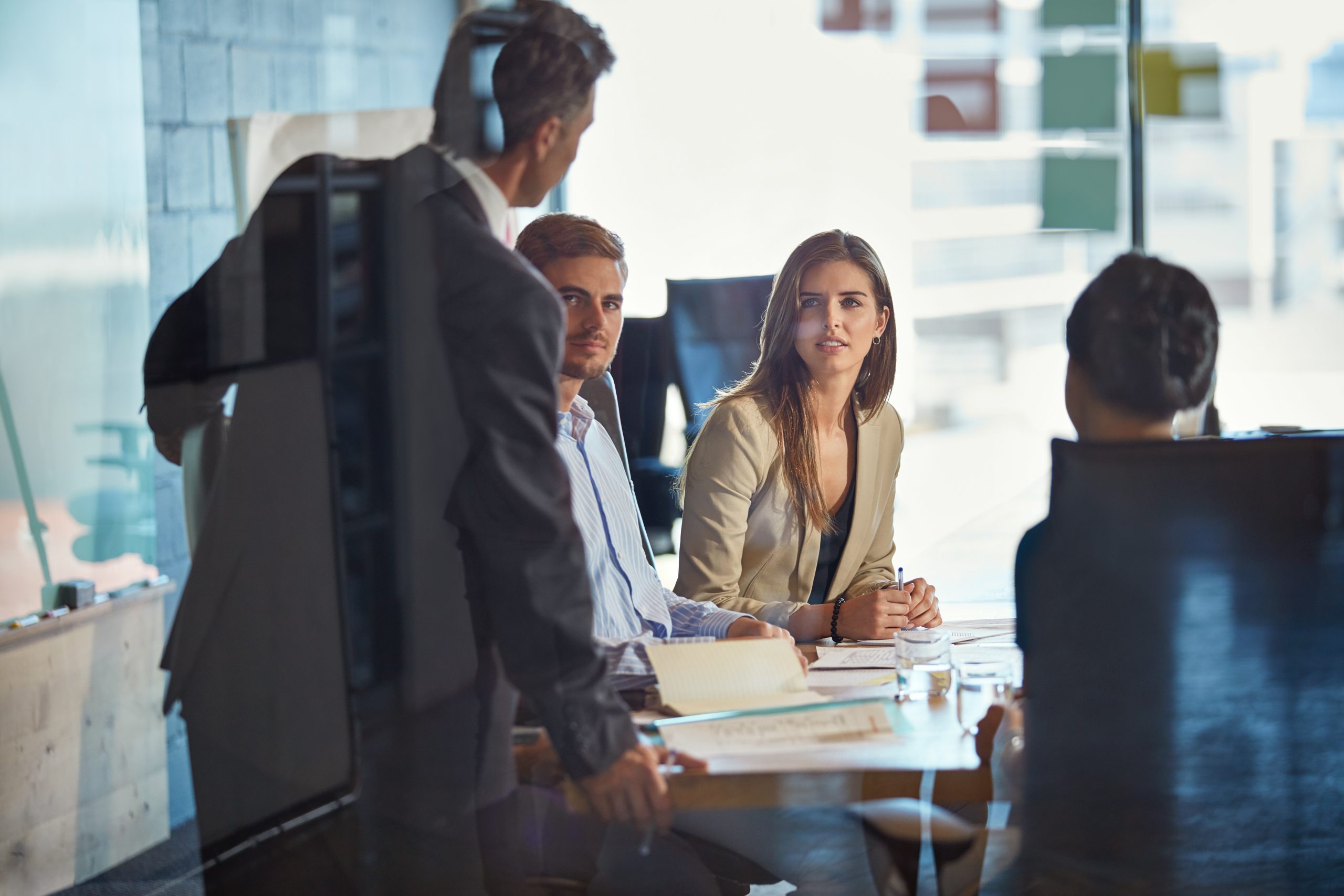 Four people in business attire sit around a conference table in an office, having a discussion with documents and glasses of water on the table.