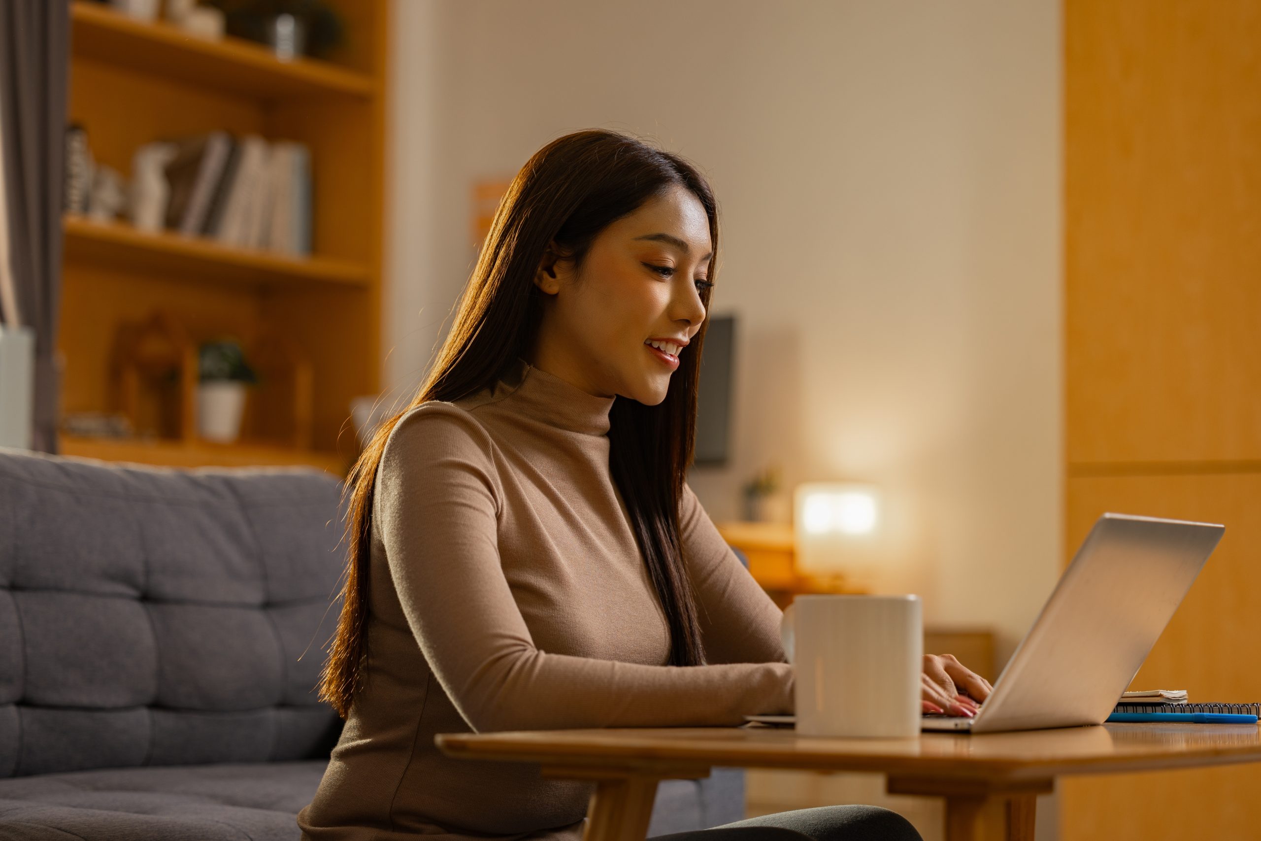 A woman sits on a couch, typing on a laptop placed on a wooden table with a white mug, in a warmly lit living room.